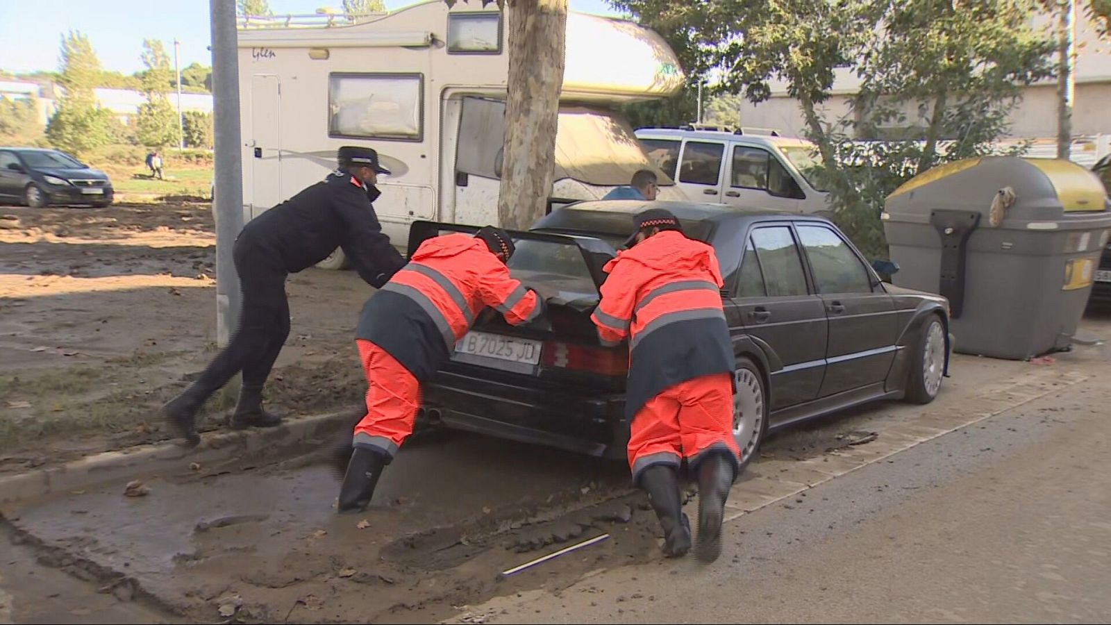 Tres efectivos de emergencias y un agente empujan un coche negro clásico para sacarlo del fango en una calle afectada por inundaciones.