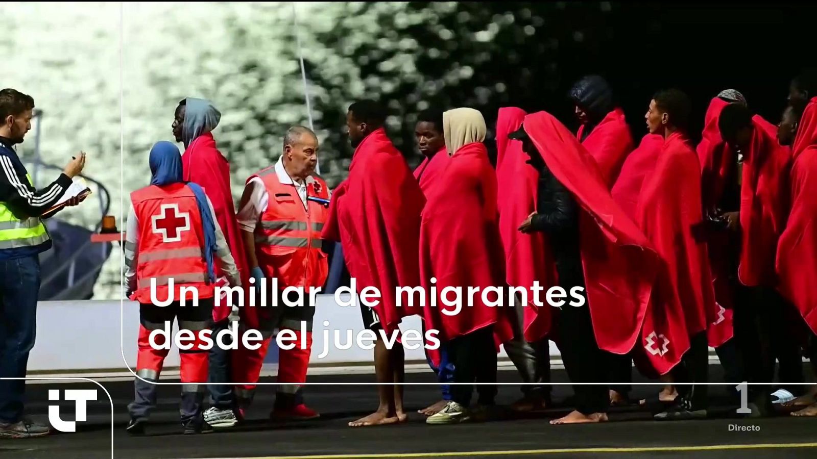 Grupo de personas migrantes en un muelle cubiertas con mantas térmicas rojas y siendo atendidas por voluntarios de la Cruz Roja en un puerto.