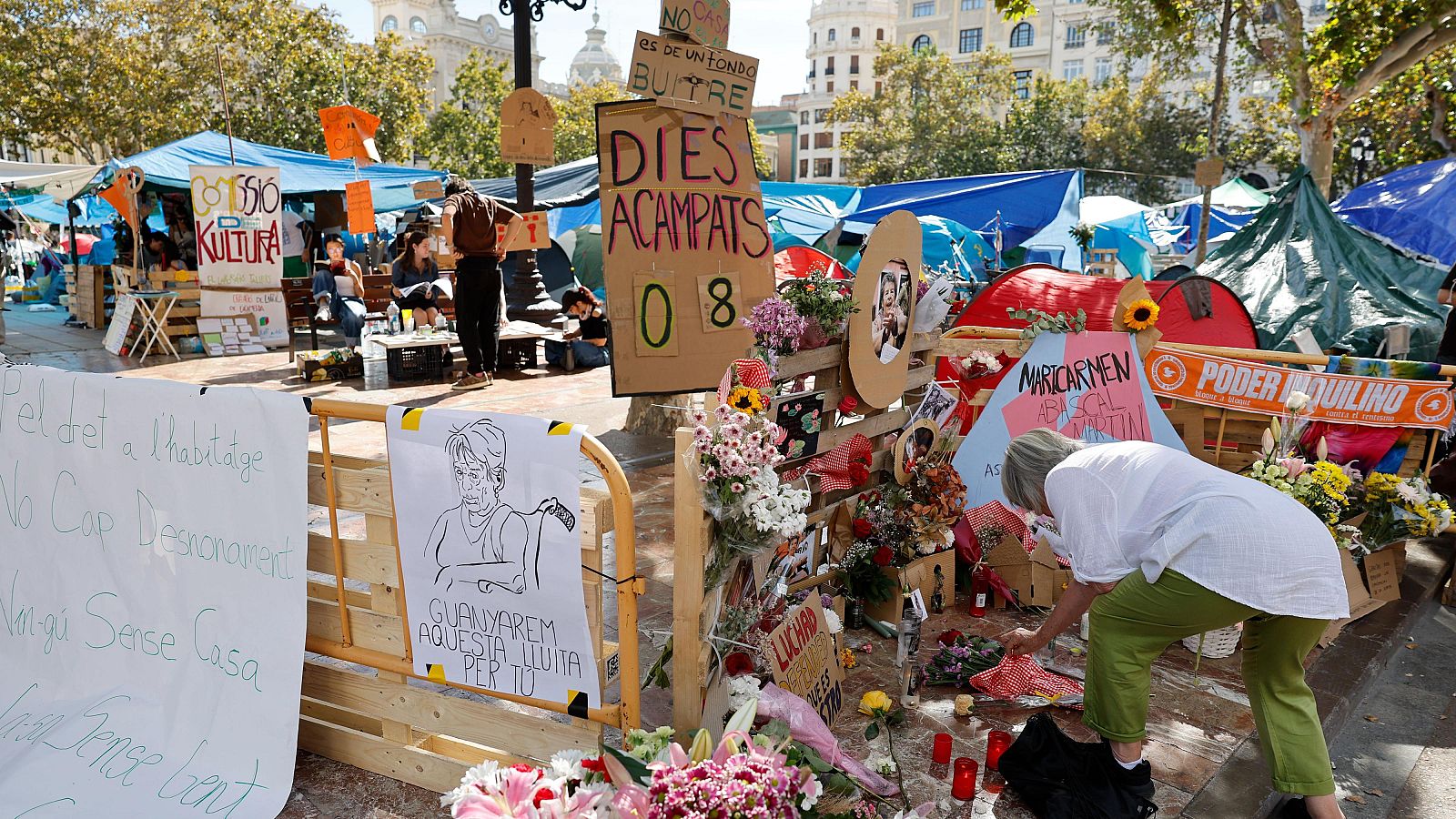 Campamento de protesta con tiendas, pancartas sobre vivienda y un altar conmemorativo con velas y flores.