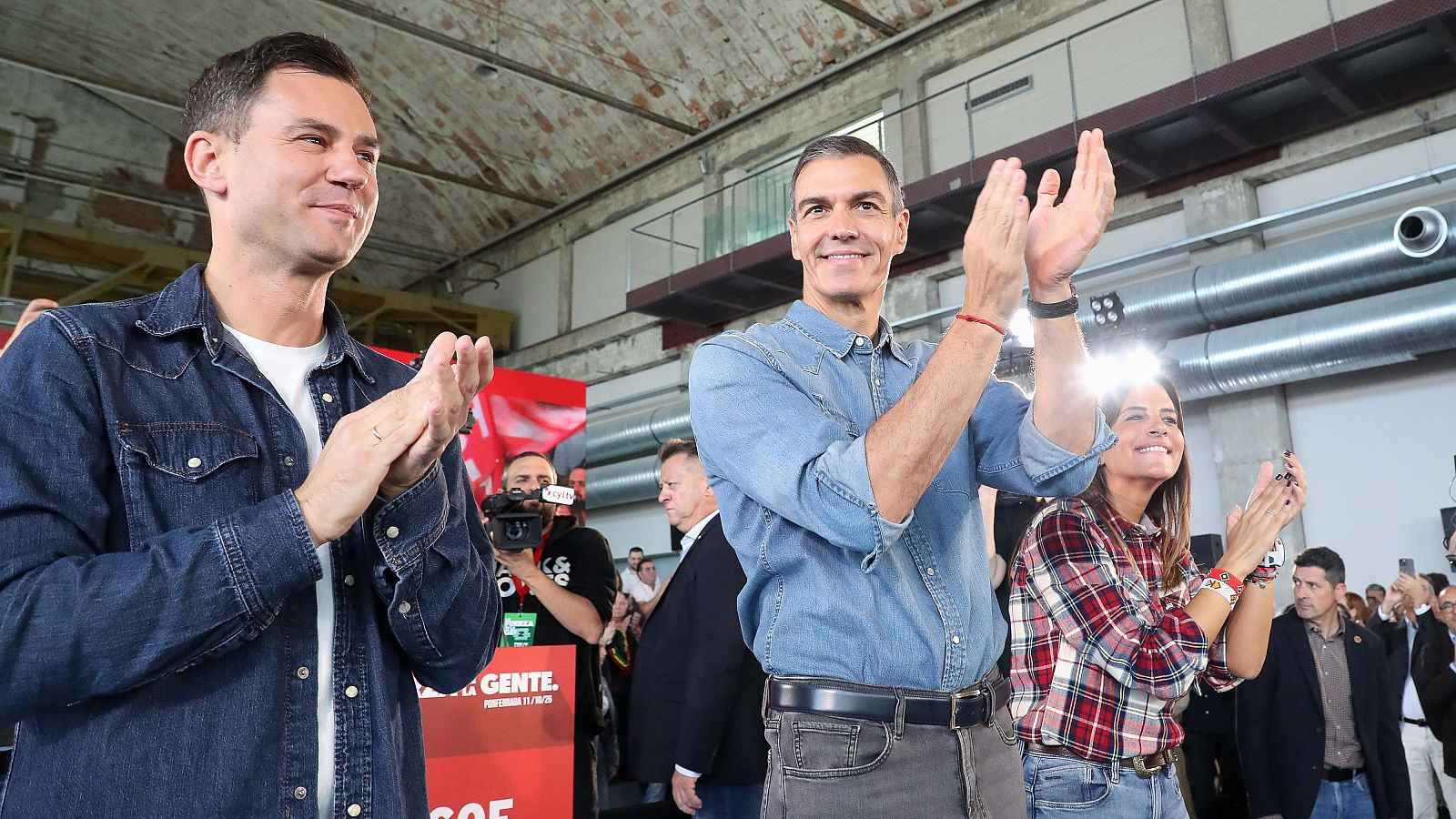 Tres políticos aplauden sonrientes en un acto público en una nave industrial con elementos metálicos visibles.