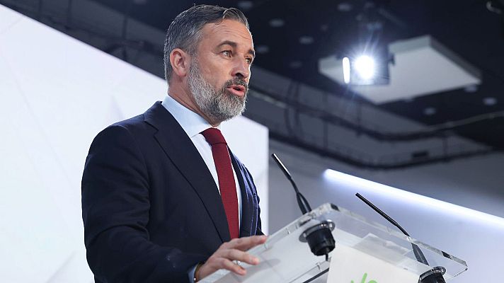 El político Santiago Abascal comparece ante un atril transparente luciendo traje azul marino y corbata roja con gesto serio.