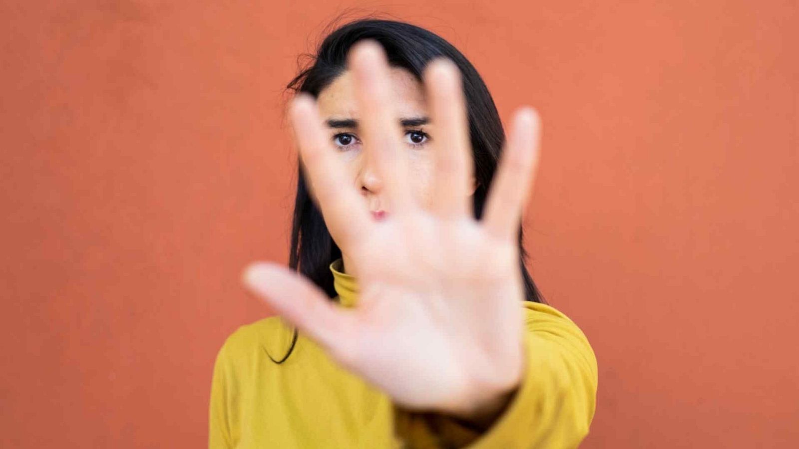 Mujer joven con jersey amarillo mostaza muestra la palma de la mano hacia la cámara en gesto de alto, con la mirada seria fija en el espectador sobre fondo terracota.
