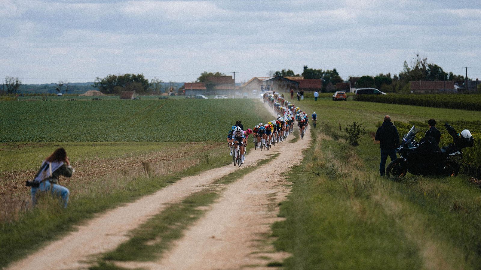 Un pelotón de ciclistas avanza por un camino de tierra rural bordeado de campos verdes, observado por varios espectadores y personal del evento.