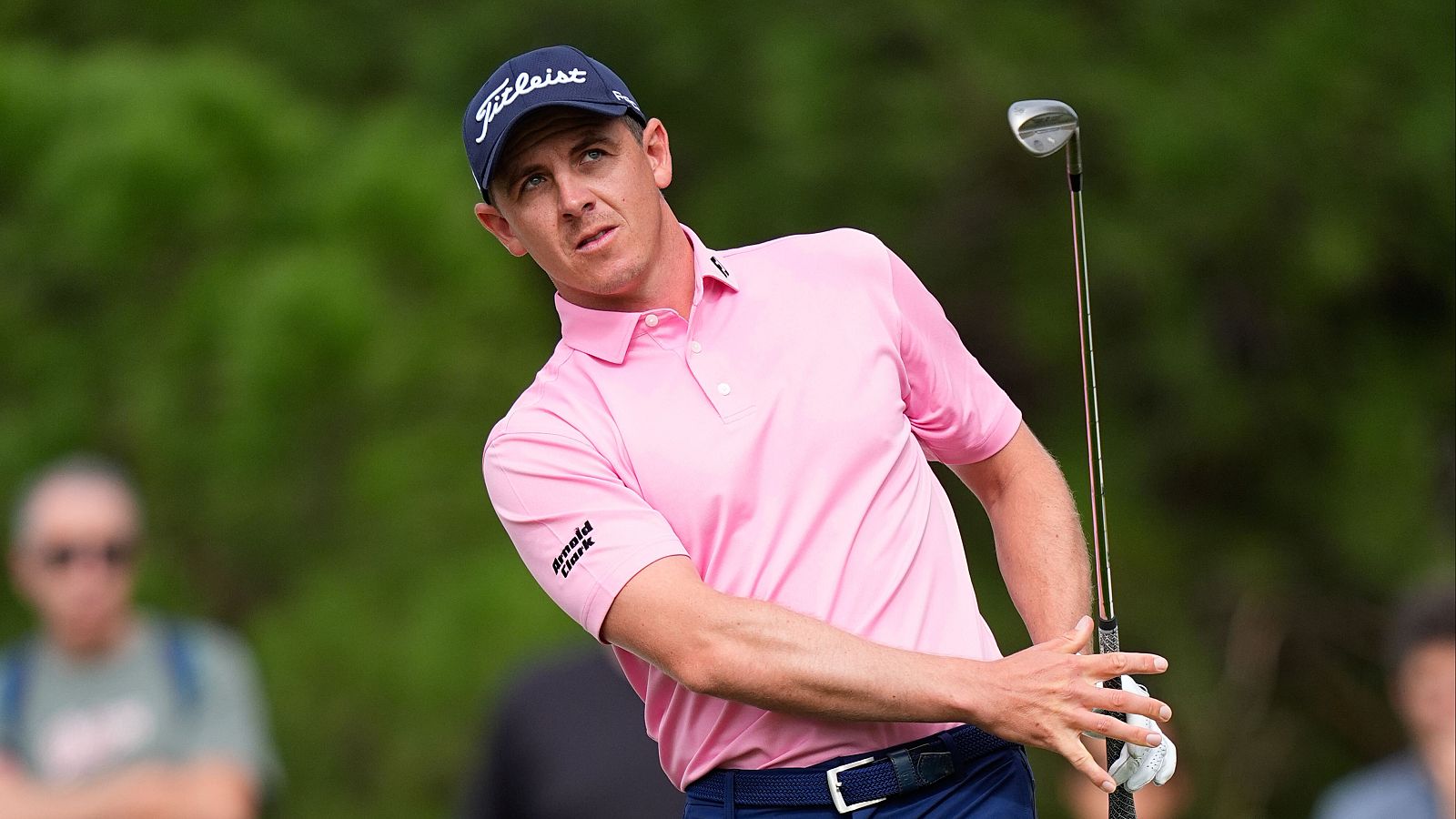 Plano medio del golfista Grant Forrest en un campo de golf, vistiendo polo rosa y gorra azul, sosteniendo un palo mientras observa el golpe.