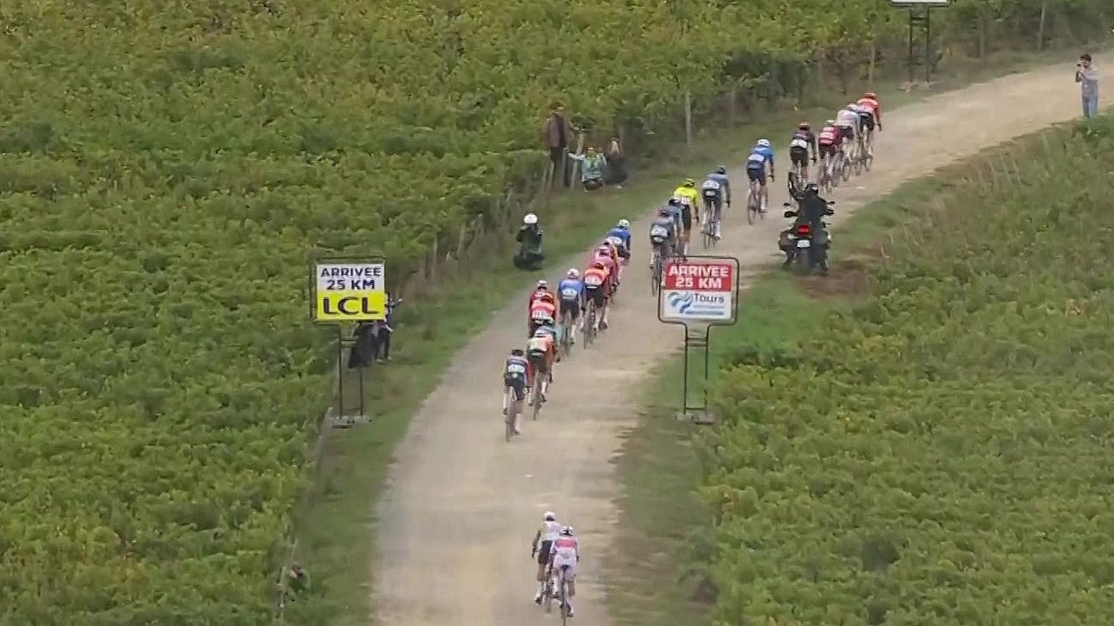 Carrera ciclista profesional sobre un camino de tierra entre viñedos con carteles que indican la meta a 25 km.