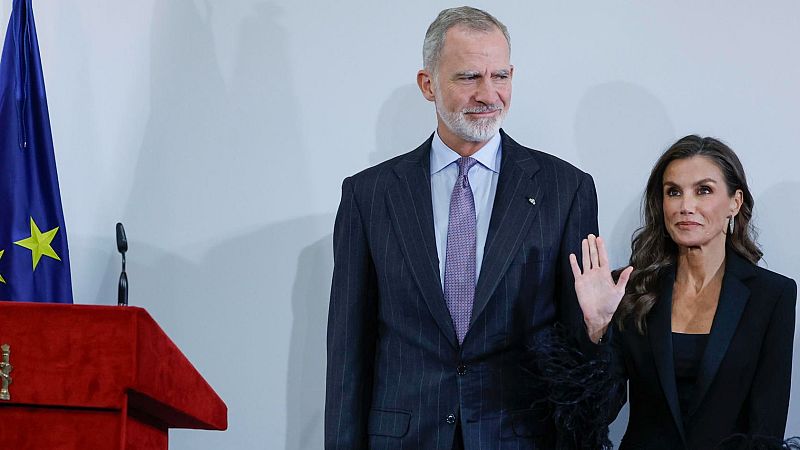 El Rey Felipe VI con traje formal y la Reina Letizia con conjunto negro de plumas en un acto oficial ante una bandera de la UE y un atril.