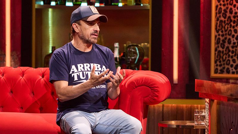 José Mota, sentado en un sofá rojo y vestido de forma casual, gesticula con las manos en un plató con iluminación cálida.