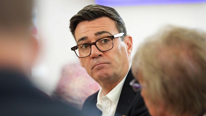El político británico Andrew Burnham, con gafas y expresión seria, aparece en primer plano durante una reunión pública con traje oscuro.