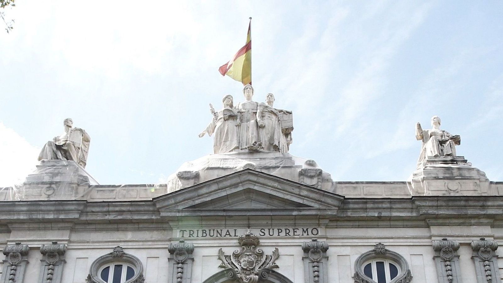 Plano contrapicado de la fachada superior del Tribunal Supremo en Madrid, con esculturas, el escudo y la bandera de España.