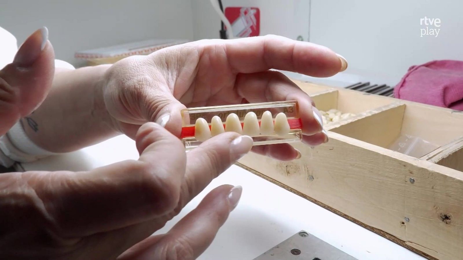Manos de una técnico con manicura y tatuaje examinan una guía transparente con dientes artificiales frontales sobre una mesa de laboratorio.