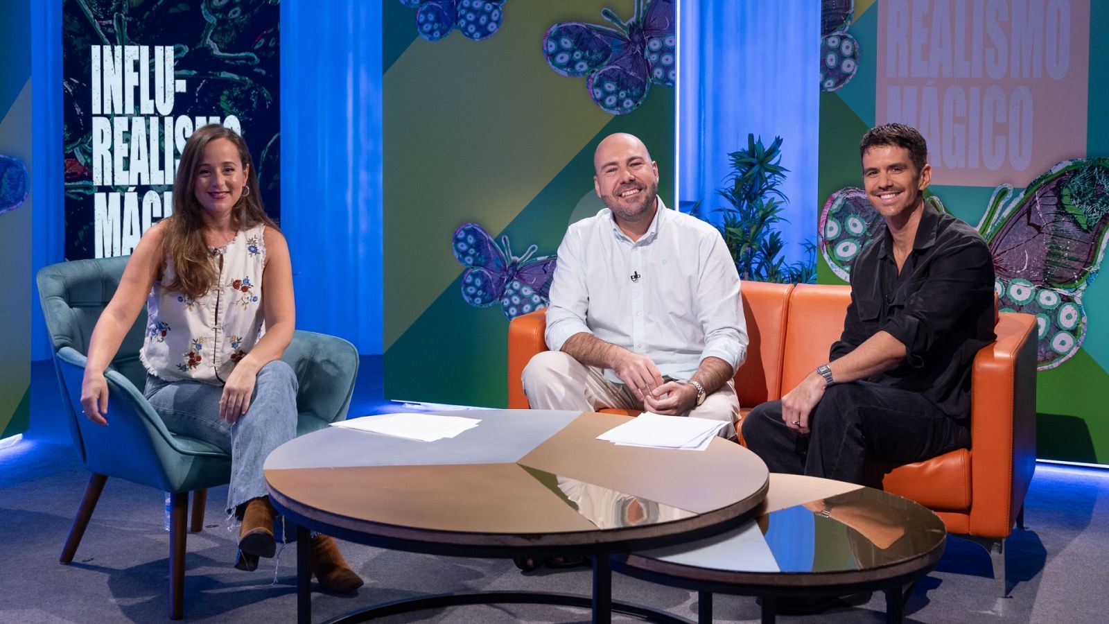 Plató de televisión con tres personas sonrientes sentadas en butacas y un sofá naranja frente a mesas bajas y un fondo decorado con paneles iluminados.