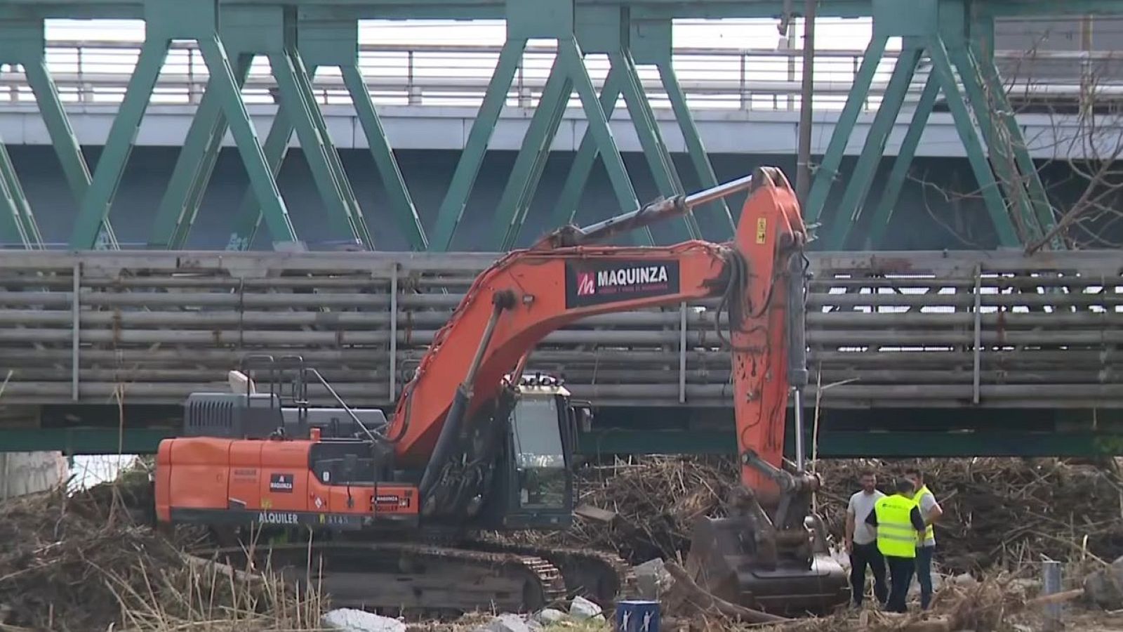 Excavadora naranja de MAQUINZA trabajando bajo un puente metálico verde junto a tres operarios en un terreno de tierra y ramas.
