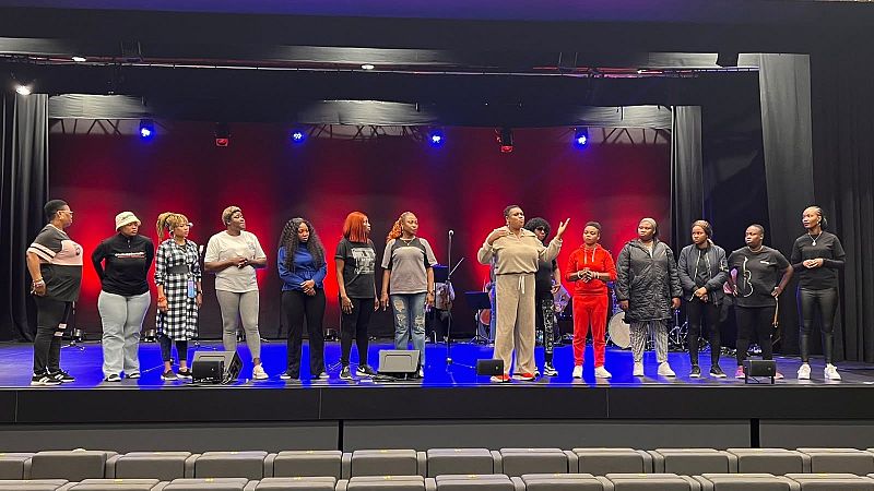 Catorce mujeres ensayan en el escenario de un teatro con butacas vacías, vistiendo ropa informal y deportiva bajo luces azules y rojas.