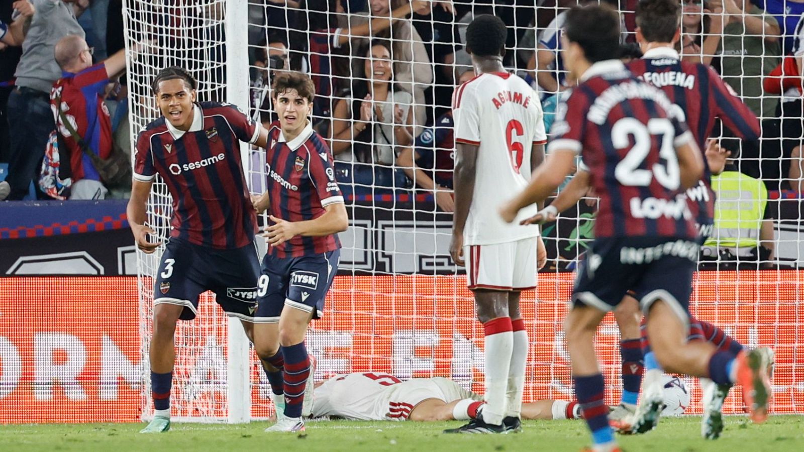 Dos futbolistas del Levante UD celebran un gol con euforia junto a la red, mientras un jugador del Alcorcón lamenta la derrota.