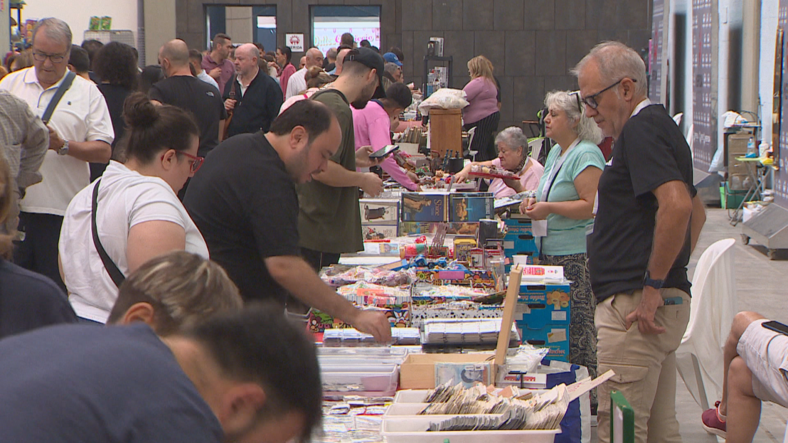 Pabellón ferial lleno de mesas con artículos de colección, donde varios asistentes observan y examinan diversos objetos expuestos para su venta.