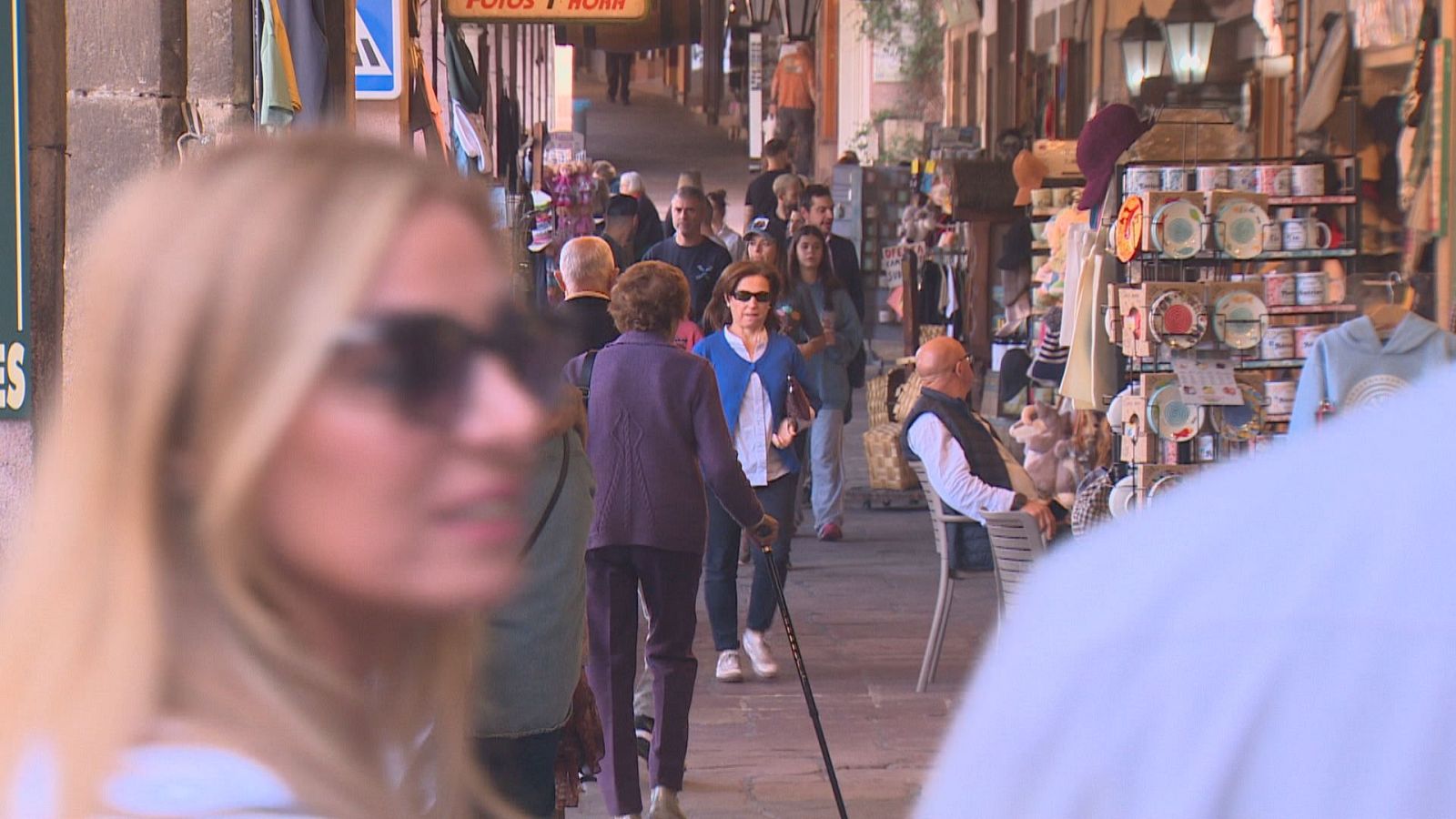 Calle peatonal soportalada y concurrida con turistas paseando, tiendas de recuerdos y personas en primer plano.