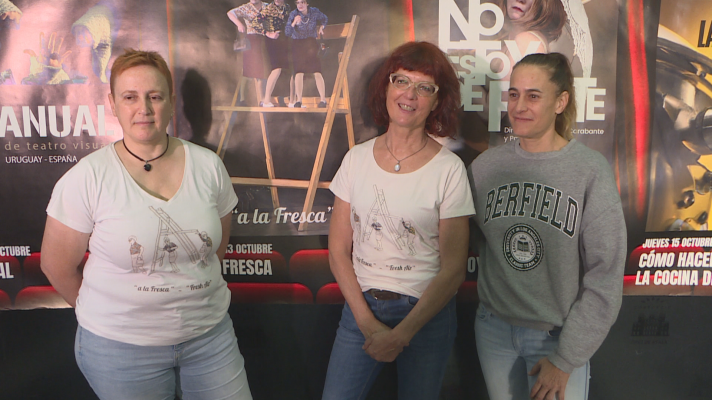Tres mujeres posan en un photocall de un festival de teatro frente a carteles de obras, vistiendo dos de ellas camisetas con la inscripción «a la fresca» y otra una sudadera gris.