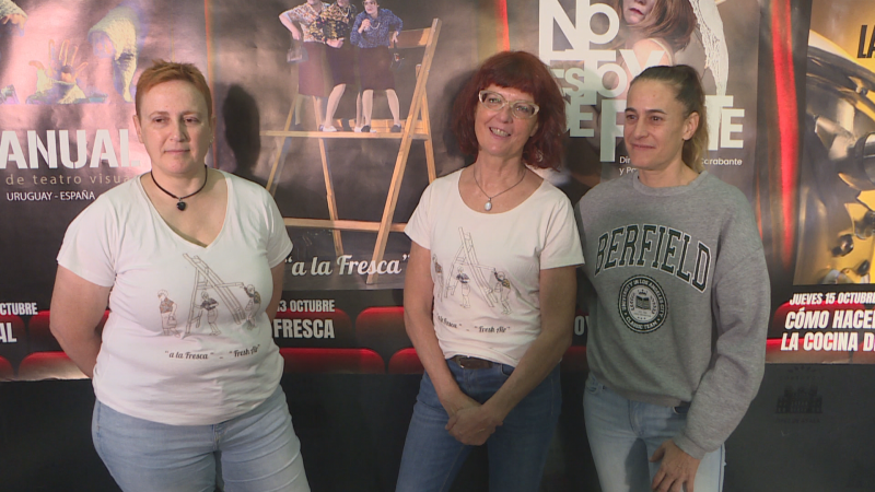 Tres mujeres posan en un photocall de un festival de teatro frente a carteles de obras, vistiendo dos de ellas camisetas con la inscripción «a la fresca» y otra una sudadera gris.