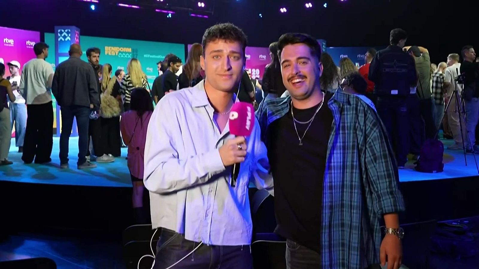 Dos jóvenes en un evento de prensa ante un photocall iluminado; uno sostiene un micrófono con el logo de RTVE Play y el otro sonríe con una sobrecamisa a cuadros.