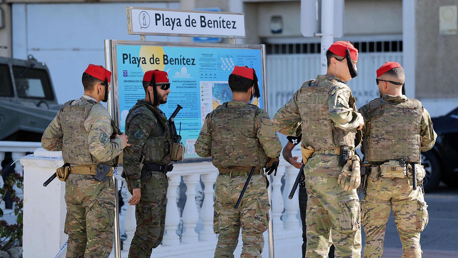 Cinco militares españoles de Regulares con uniforme de camuflaje y fez rojo se encuentran junto a un panel de la Playa de Benítez en una zona costera urbana.