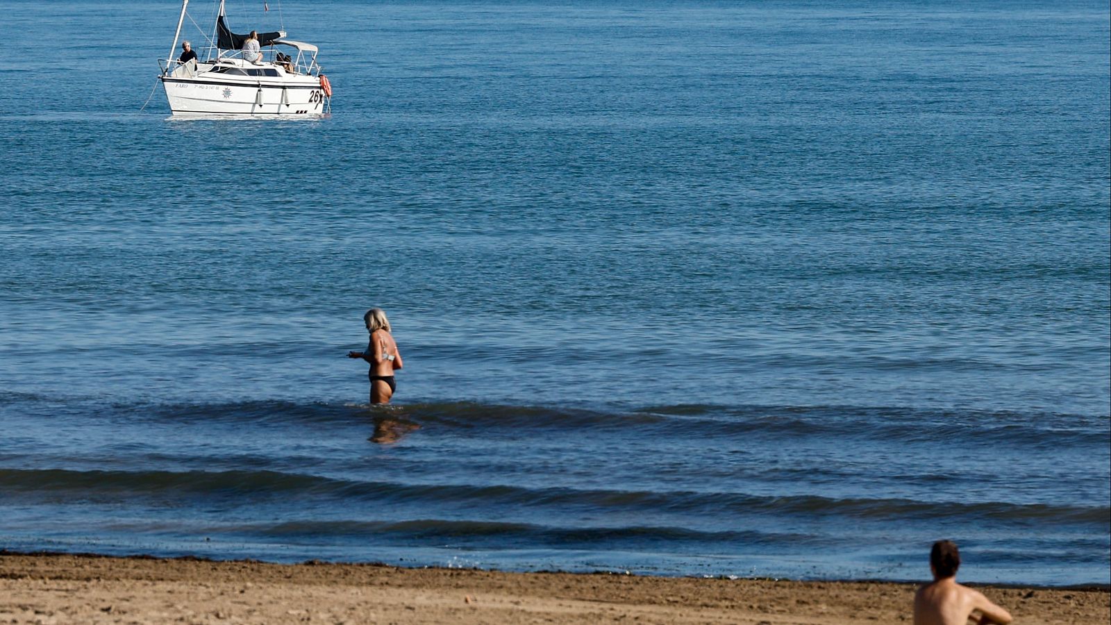 Escena costera veraniega con una mujer caminando por el mar y un pequeño velero blanco flotando a lo lejos bajo una luz diurna.