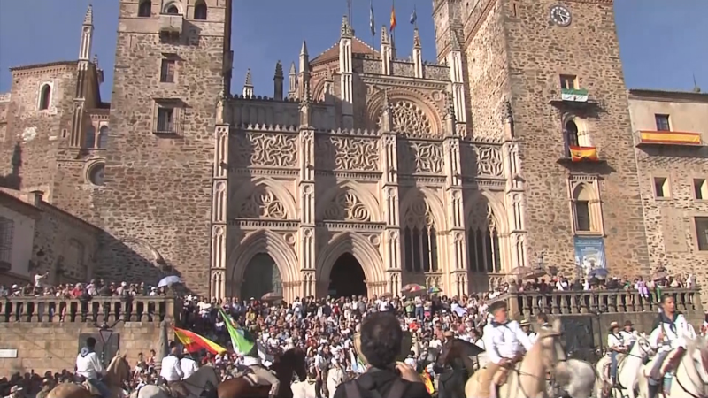 Vista frontal del Real Monasterio de Guadalupe durante una festividad al aire libre con multitud de peregrinos y jinetes a caballo en la plaza.