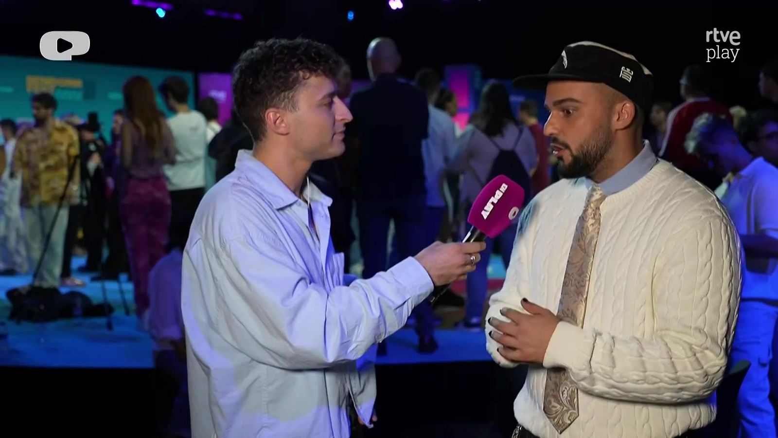 Un reportero con camisa azul entrevista a Sev's, quien lleva gorra negra, jersey blanco y corbata, en un set con iluminación rosa y fría.