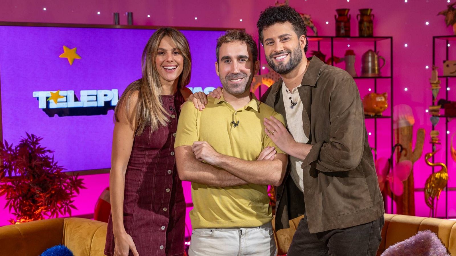 Tres presentadores posan sonrientes en un plató con iluminación fucsia y morada tras un sofá mostaza, flanqueando al presentador central Aitor Albizua.