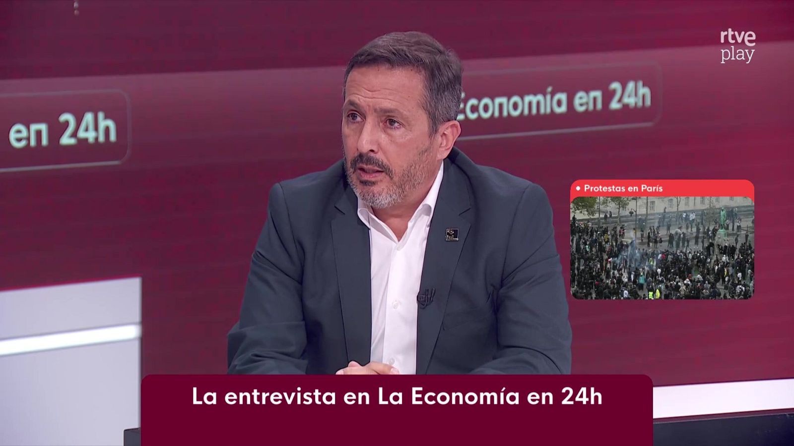 Un hombre de mediana edad es entrevistado en un plató de televisión con rótulos informativos y una ventana superpuesta sobre protestas.