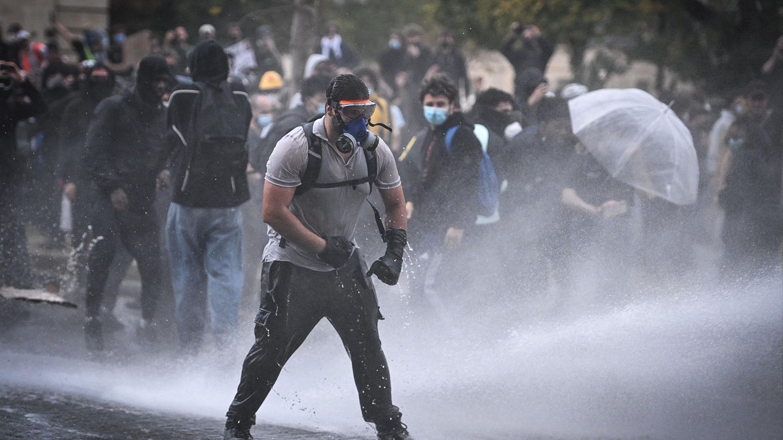 Un joven manifestante con máscara antigás y guantes negros se enfrenta desafiante mientras un chorro de agua a presión cruza la calle durante una protesta.