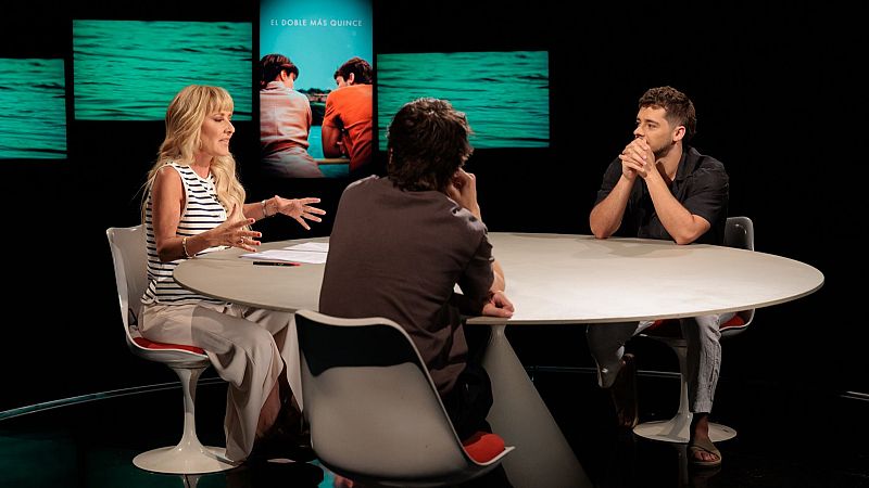 Plató de televisión con tres personas conversando alrededor de una mesa blanca sobre fondo negro y pantallas con imágenes.