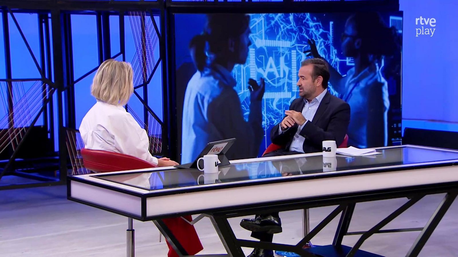Dos personas conversan sentadas en taburetes rojos alrededor de una mesa alta en un plató con un videowall azul de temática tecnológica al fondo.