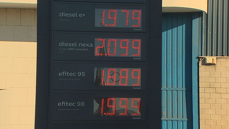 Monolito digital vertical de precios de carburantes con dígitos LED rojos y fachada con portón azul de una gasolinera.