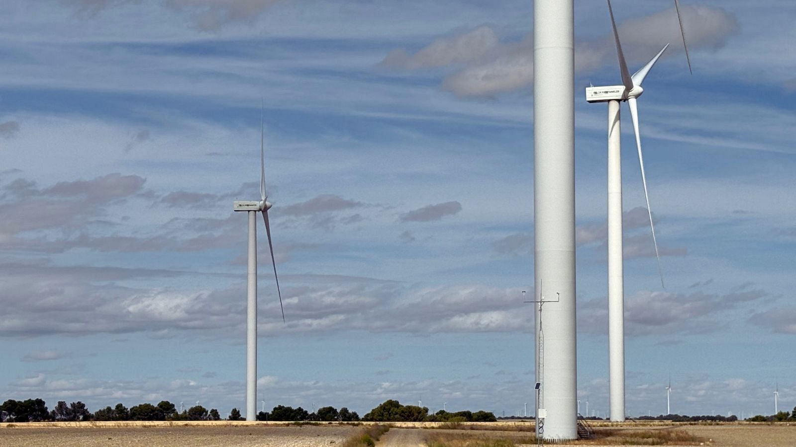 Vista panorámica diurna de un parque eólico en una llanura rural con aerogeneradores blancos, mástil de medición y campos agrícolas secos bajo un cielo azul con nubes finas.