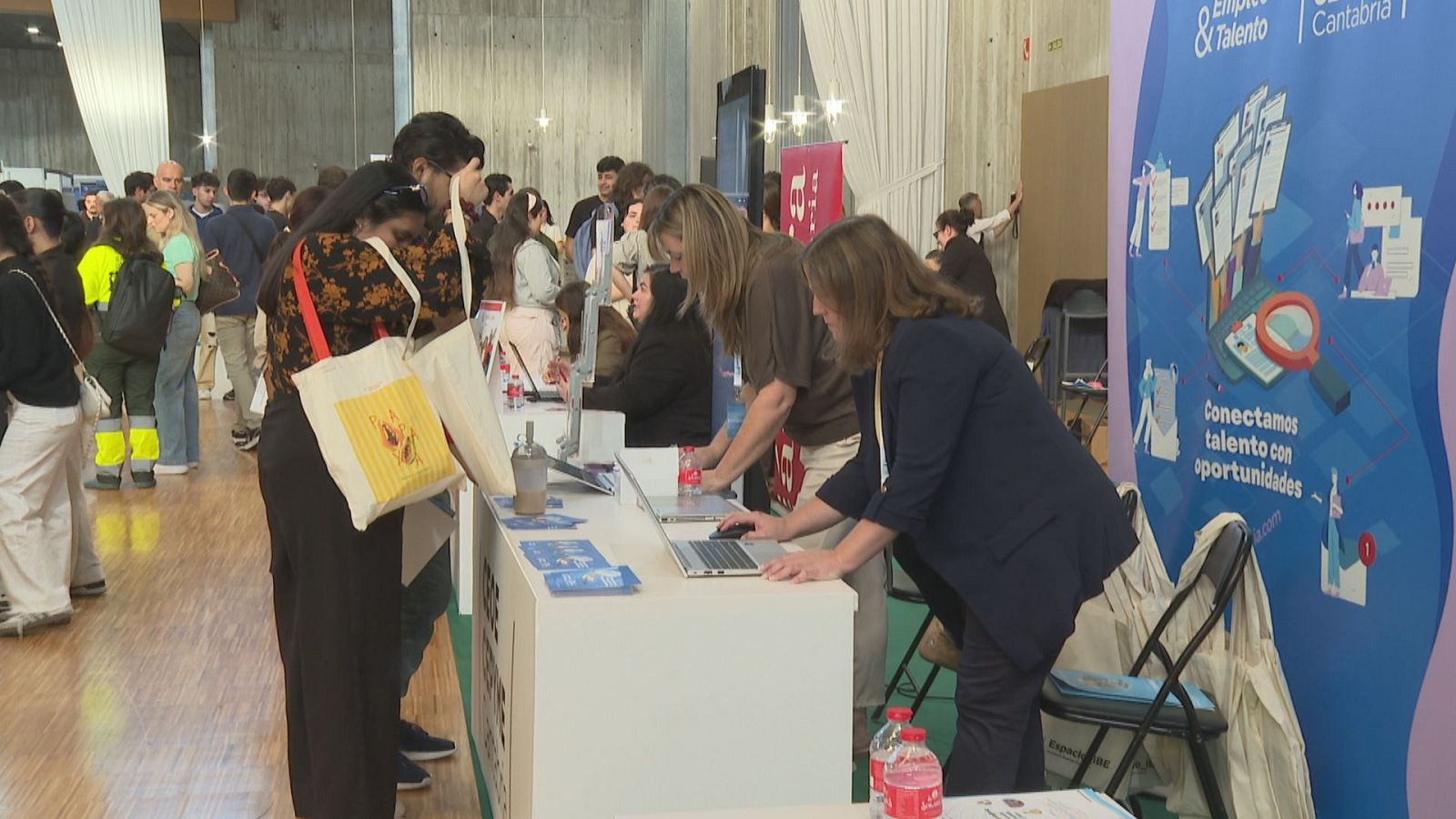 Feria de empleo en un recinto interior concurrido, mostrando un mostrador de atención, asistentes haciendo fila y un panel enrollable azul.