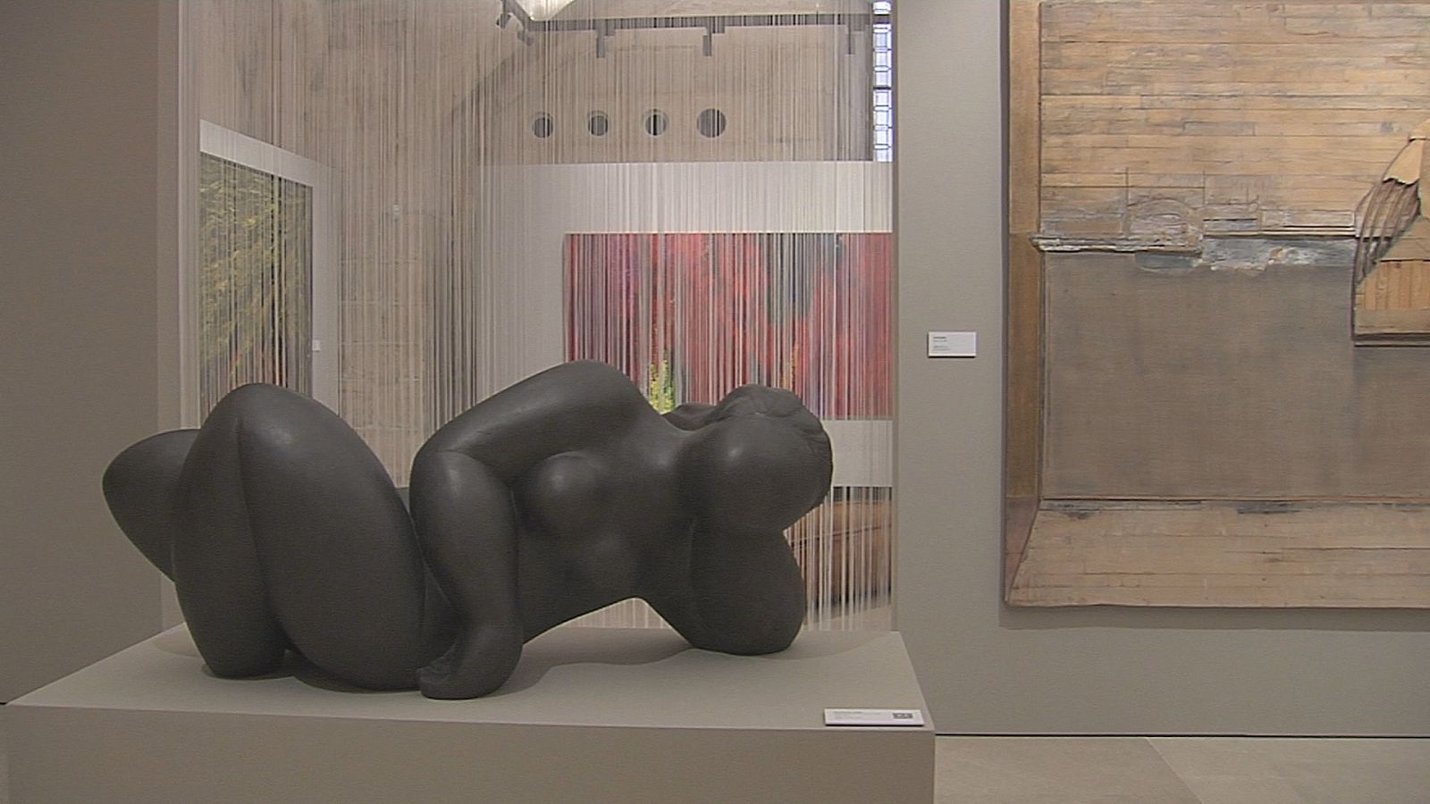 Sala de museo con una escultura de mujer reclinada sobre una peana gris, lienzos en las paredes y una instalación translúcida de hilos verticales.
