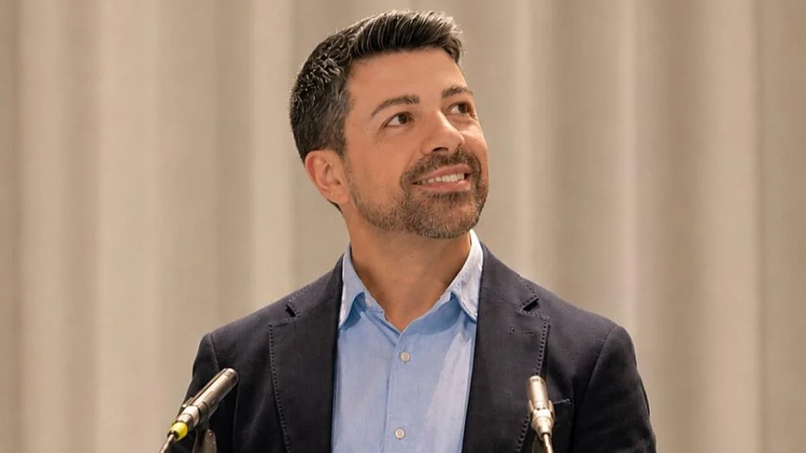 Hombre de mediana edad con barba y americana azul marino sonríe emocionado tras un atril con micrófonos.