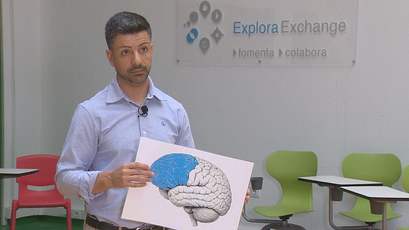 Un profesor con camisa azul sostiene una lámina con la ilustración de un cerebro en un aula moderna.