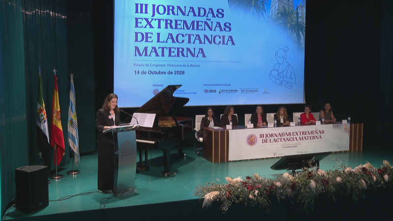 Escenario de unas jornadas con pantalla de proyección, ponente en atril, mesa presidencial con varias mujeres, banderas oficiales y arreglo floral.