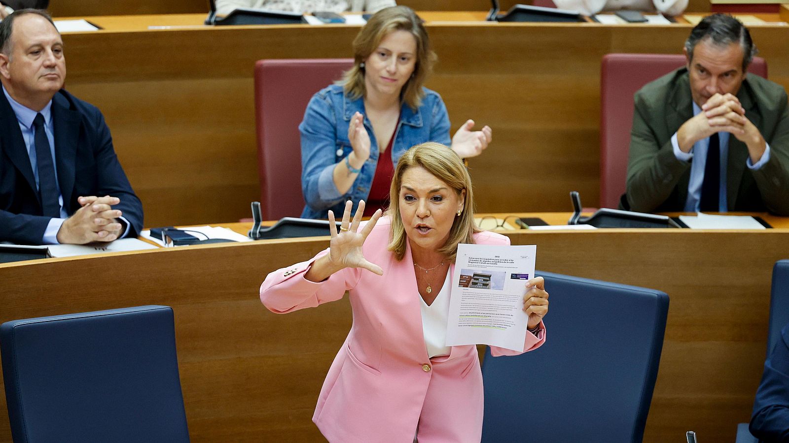 La diputada Alicia Sánchez-Camacho interviene en la Asamblea de Madrid con una chaqueta rosa pastel, mostrando un documento impreso y gesticulando con firmeza ante la cámara.