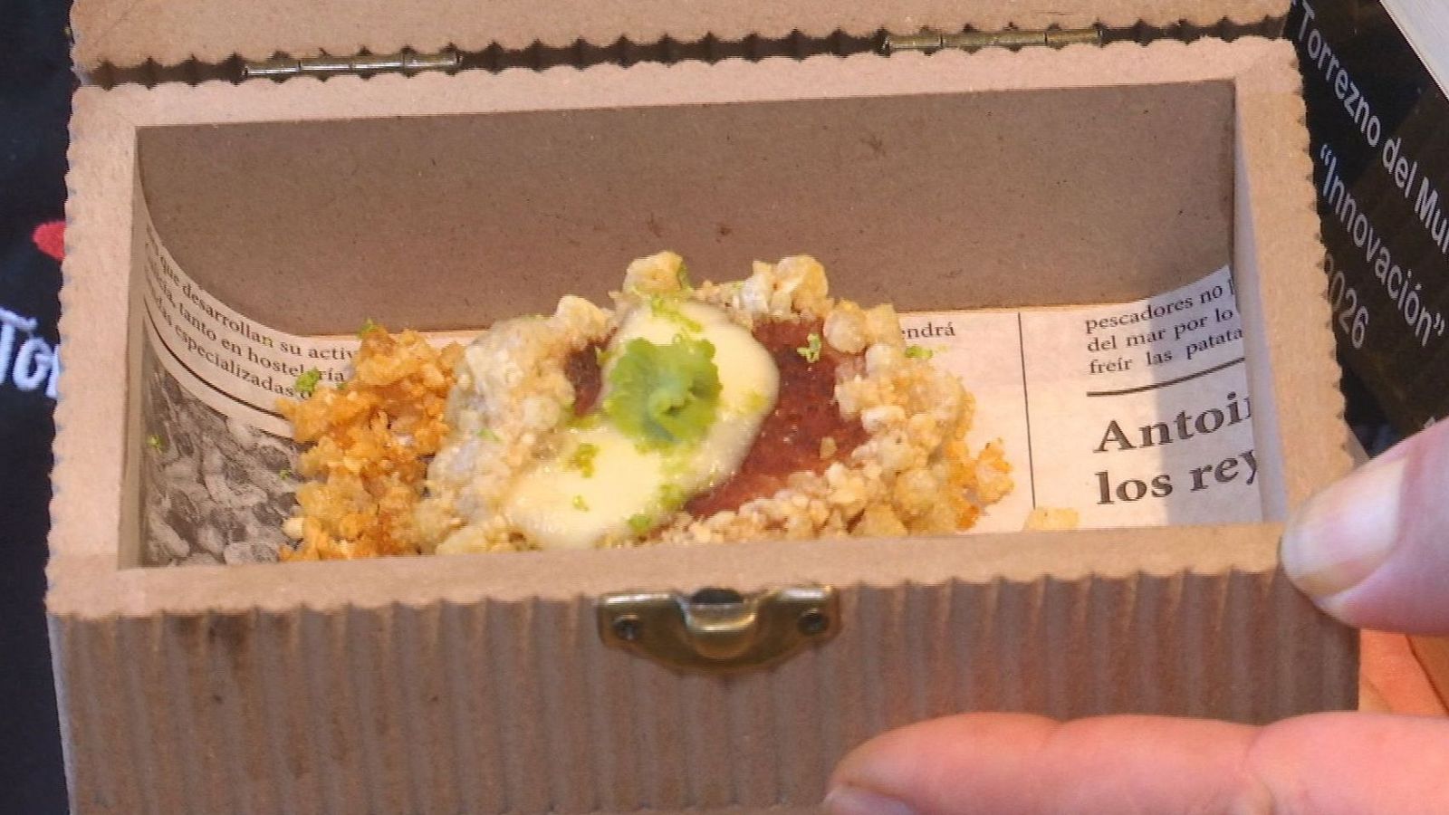 Sofisticada tapa de torrezno de vanguardia servida dentro de un pequeño cofre decorado con papel de periódico, emulsión blanca y crema verde.