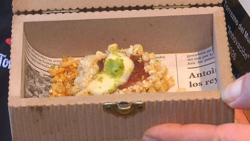 Sofisticada tapa de torrezno de vanguardia servida dentro de un pequeño cofre decorado con papel de periódico, emulsión blanca y crema verde.
