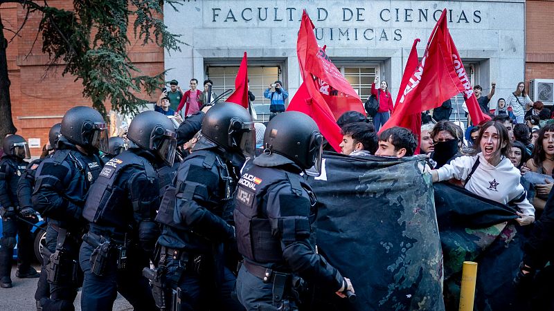 Agentes antidisturbios de la Policía Nacional y estudiantes se enfrentan frente a la entrada de un edificio universitario mientras ondean banderas rojas.