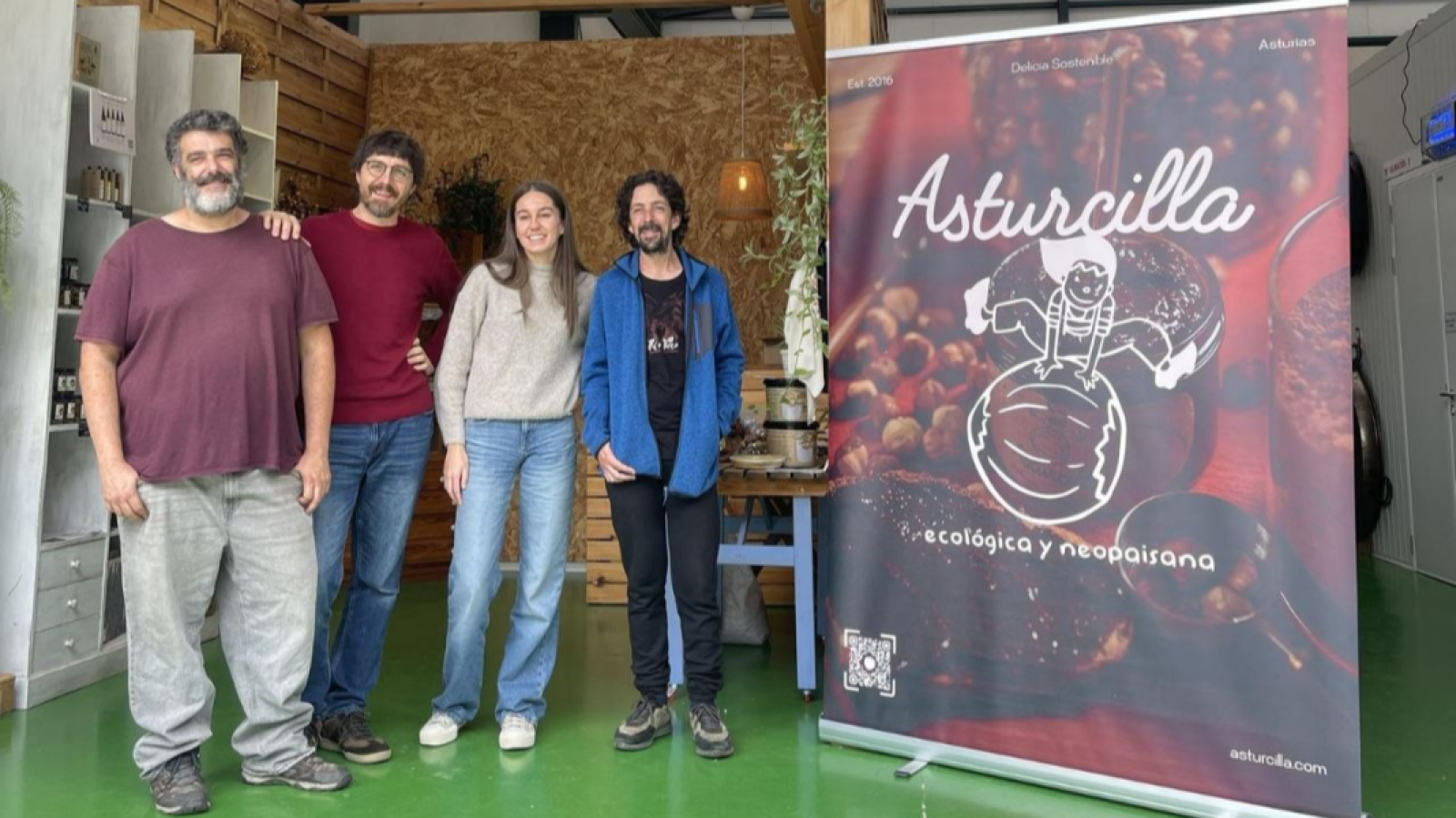 Cuatro integrantes del equipo de Asturcilla posan sonrientes junto a un cartel publicitario en su obrador artesanal.