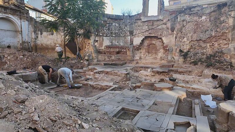 Arqueólogos trabajan en un yacimiento, excavando y limpiando restos de estructuras antiguas de piedra y ladrillo, con un pavimento de baldosas visible en primer plano.