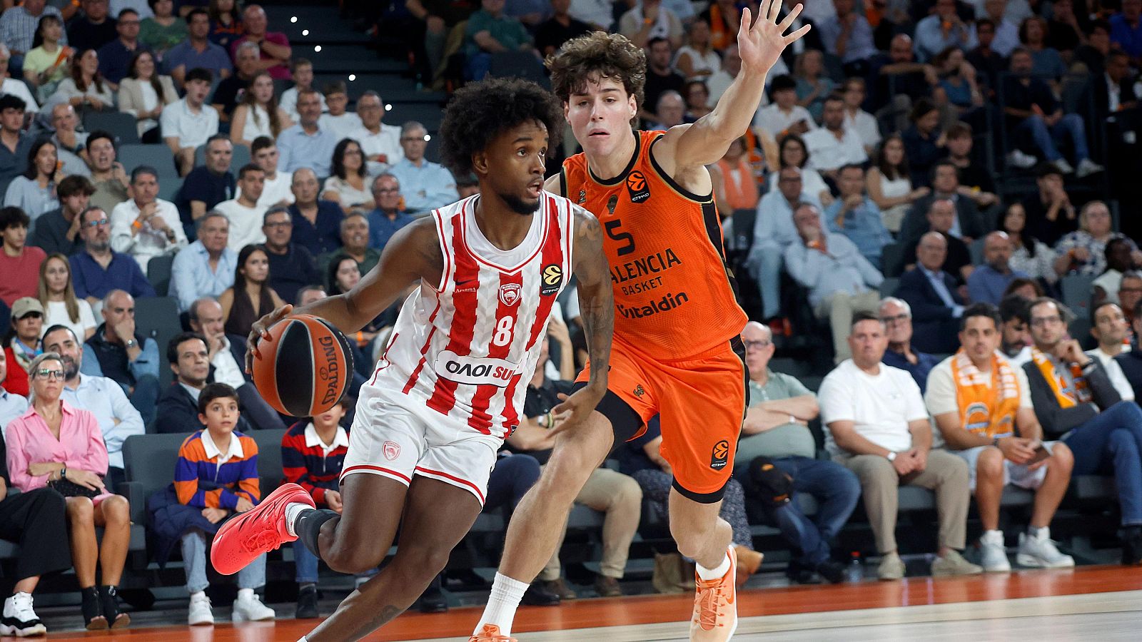 Un jugador con uniforme rojo y blanco bota el balón mientras es defendido por otro con equipación naranja en la pista de un pabellón ante las gradas llenas de espectadores.