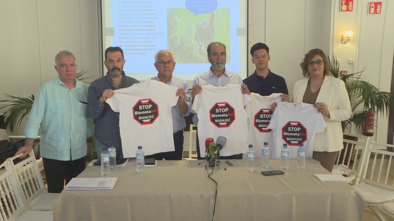 Seis pessoas de pé numa conferência de imprensa mostram camisetas brancas com o lema «STOP BiometaNO BADAJOZ» atrás de uma mesa com microfones e documentos.