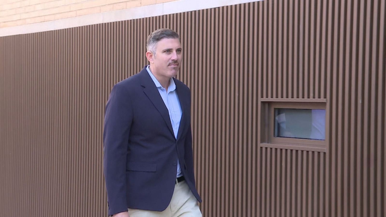 Plano medio de un hombre adulto con americana azul y camisa celeste caminando frente a un edificio moderno.