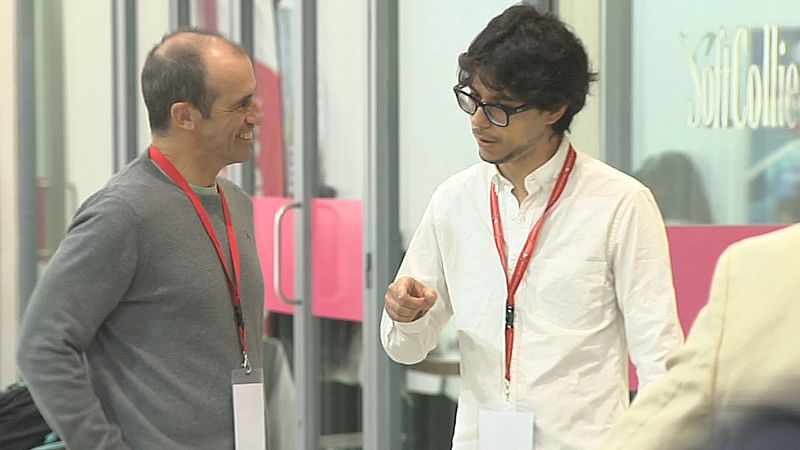 Dos hombres conversan en un evento de emprendimiento; uno de ellos gesticula explicando un proyecto mientras el otro le escucha sonriente.