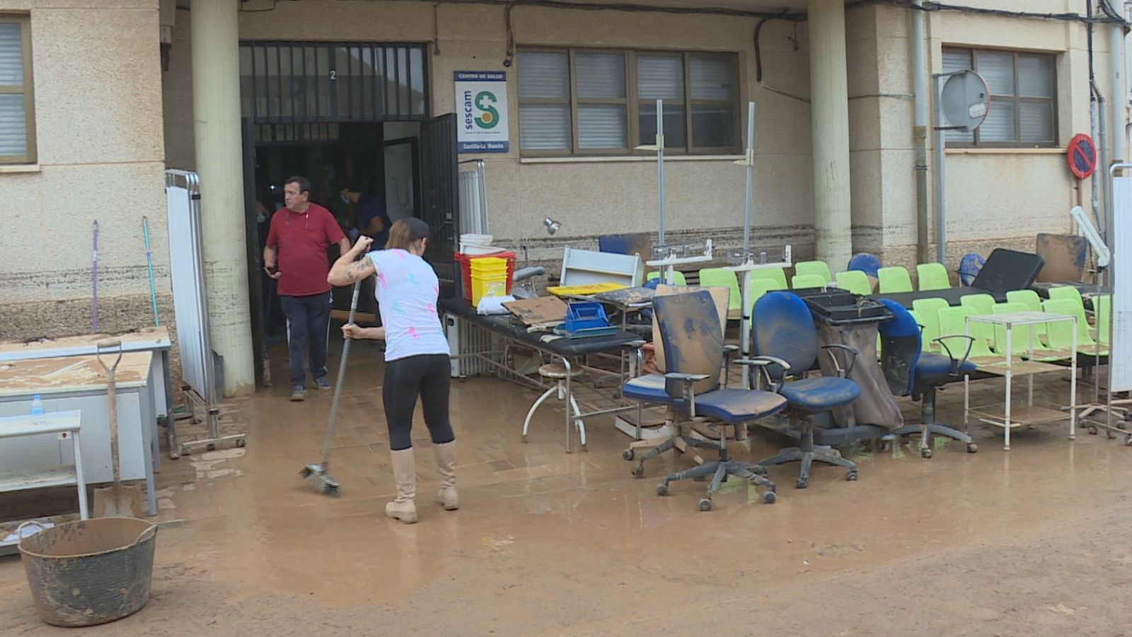Una mujer con botas de agua barre el suelo enfangado de la entrada de un centro de salud, rodeada de muebles y equipos médicos afectados por las inundaciones.
